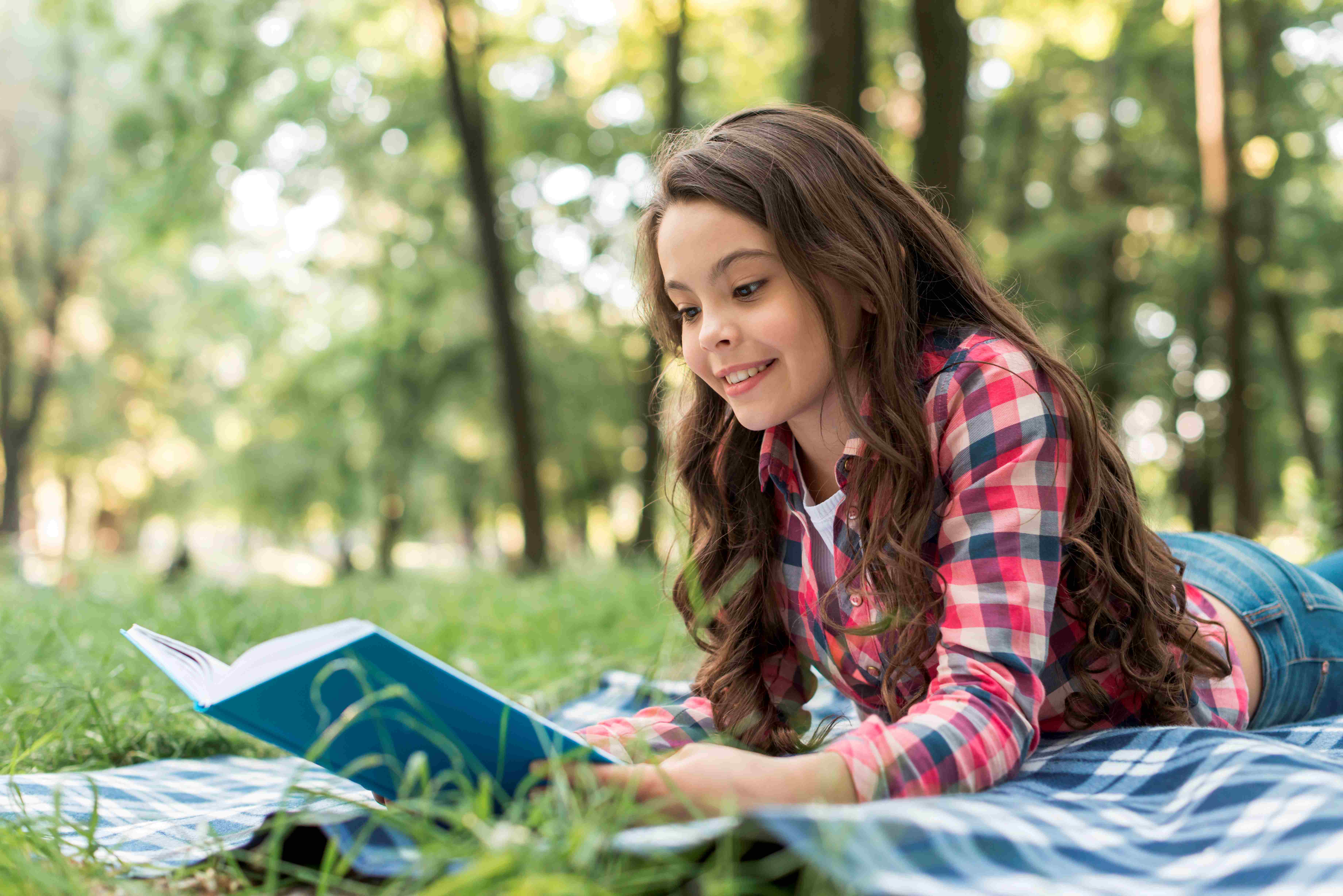 jolie fille Souriante lisant un livre en position couchée sur une couverture à carreaux au parc